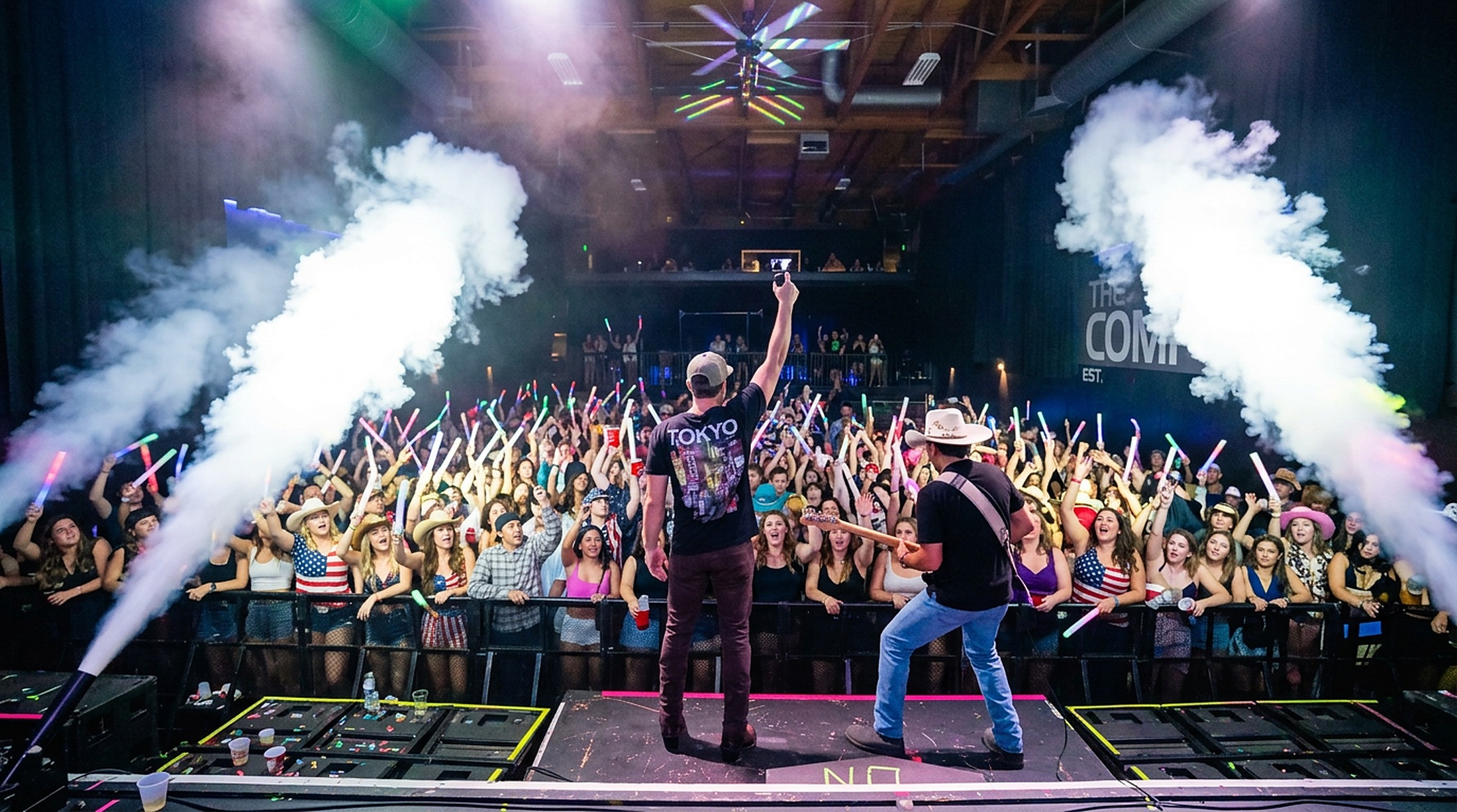 MIXMODE performing on stage — a DJ and a guitarist facing a packed crowd between two CO2 jets.